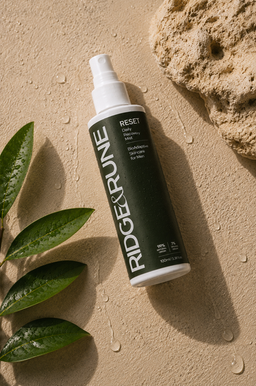 A bottle of RIDGE & RUNE RESET Daily recovery mist lies diagonally on a sandy surface with green leaves and a rock prop, casting a long shadow.